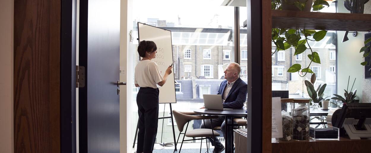 Twee personen in een vergaderzaalruimte met whiteboard. Een wat oudere man aan tafel en een vrouw van middelbare leeftijd naast het whiteboard.