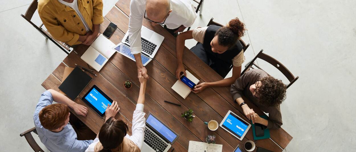 Verschillende mensen aan tafel. Iedereen heeft een elektronisch apparaat voor zich. Twee mensen in het midden geven elkaar een hand.